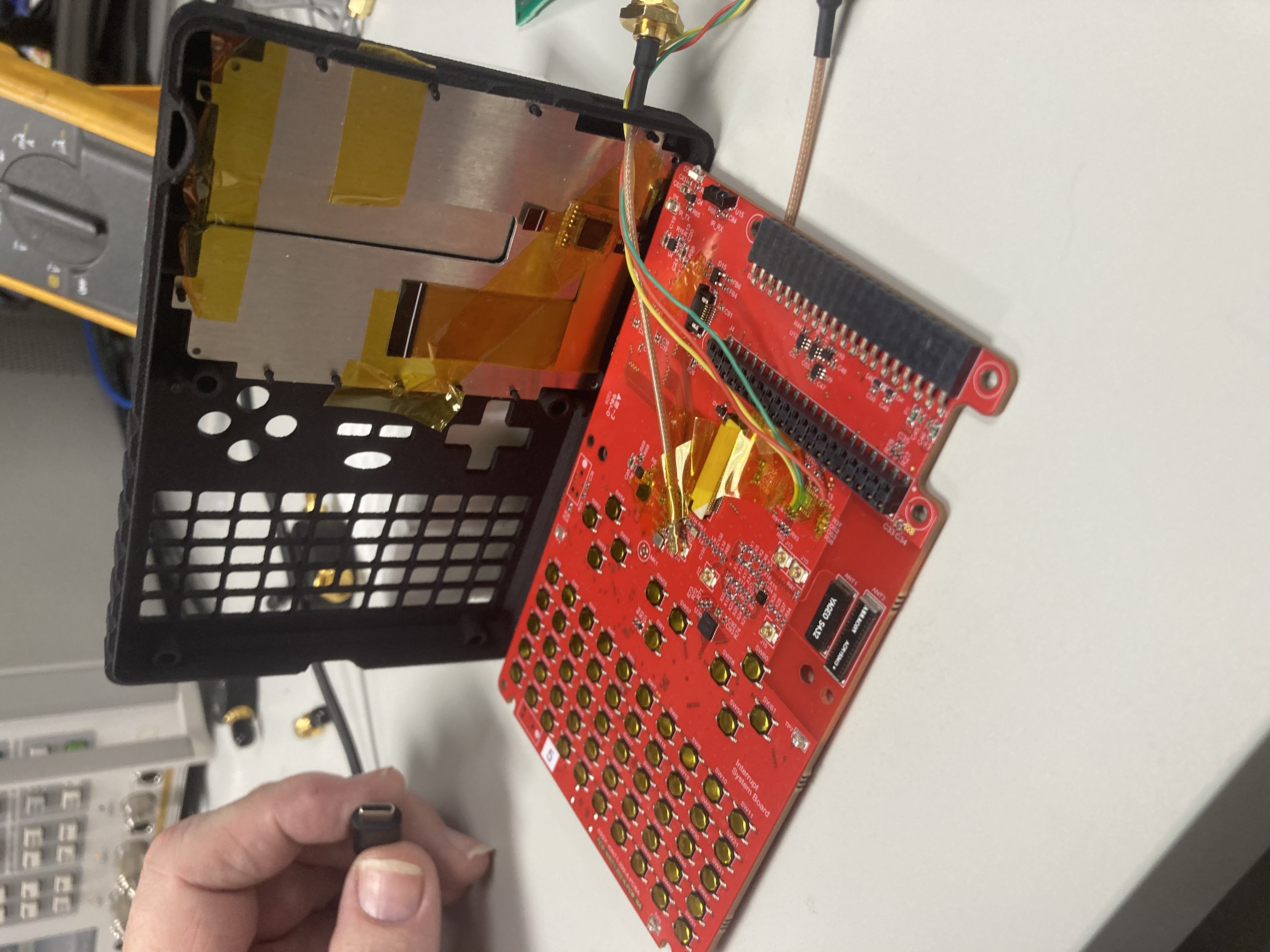 The handheld opened along its hinge: the red CC1101 system board lies flat on the bench, the back of the LCD display sits in the lifted case half, and two thin coax probes are taped down to the board near the matching-network region for VNA measurement. The chip antennas are visible in the upper corner of the red PCB, adjacent to the back of the display — the source of the antenna detuning the matching networks have to correct.