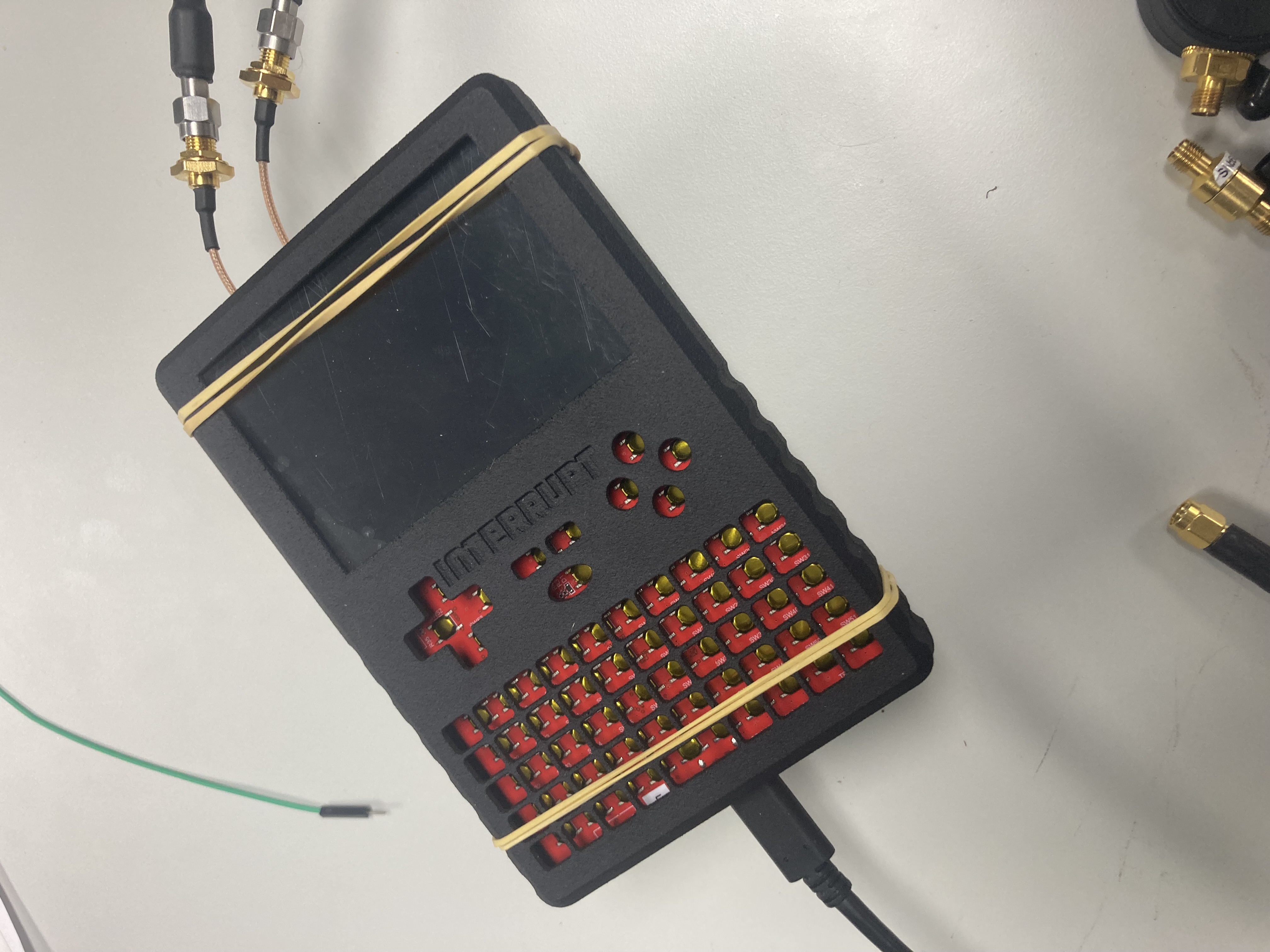 The assembled INTERRUPT handheld with display installed, rubber-banded together, USB charging cable attached, and two RF coax cables connected on the right side for in-case |S11| measurement. The red CC1101 system board is visible through the keypad cutouts on the front.